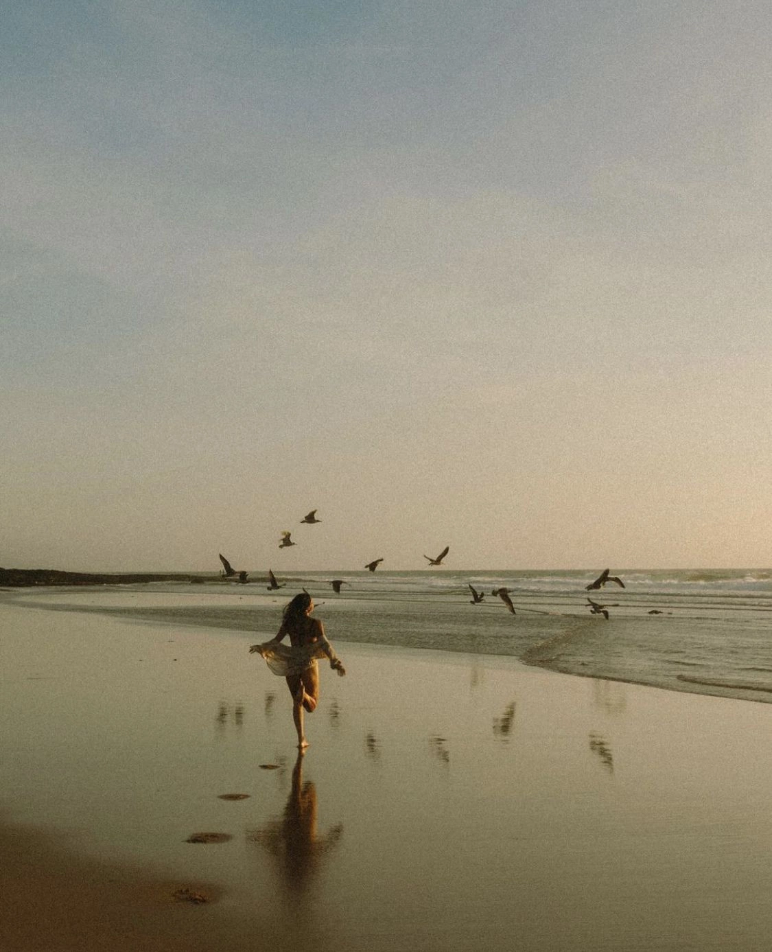 A woman running barefoot across a wet beach toward the sea, a flock of birds lifting ahead of her