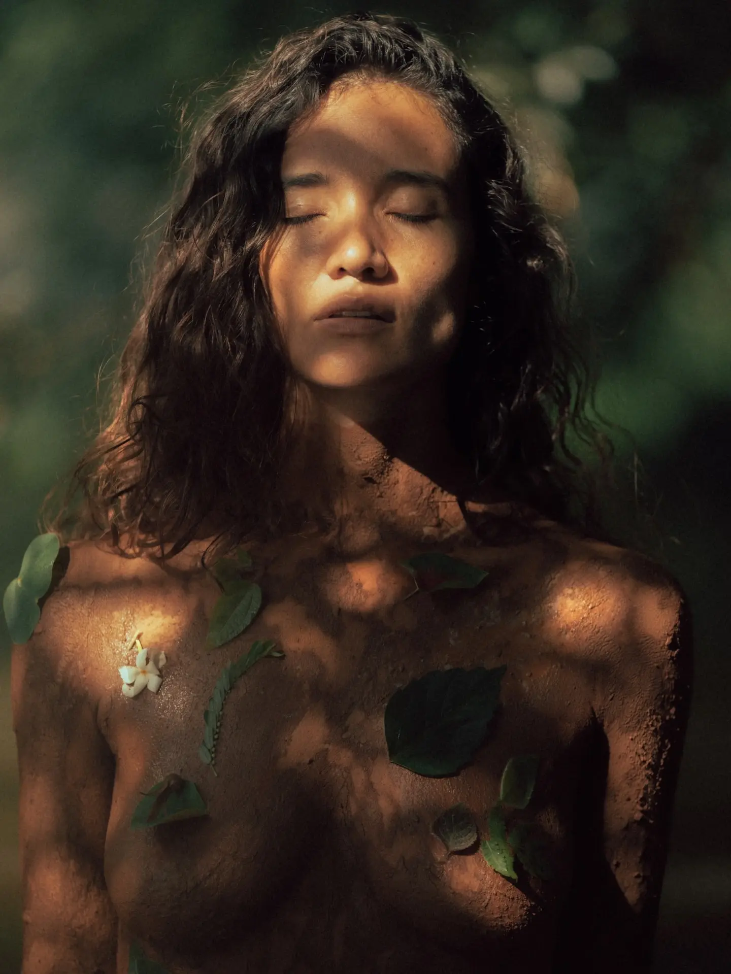 A woman with eyes closed, skin touched by earth, leaves, and soft light