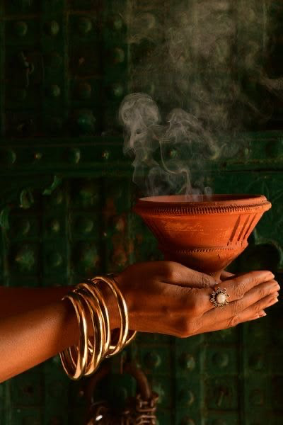 Hands with gold bangles holding a smoking clay incense bowl before a green temple door