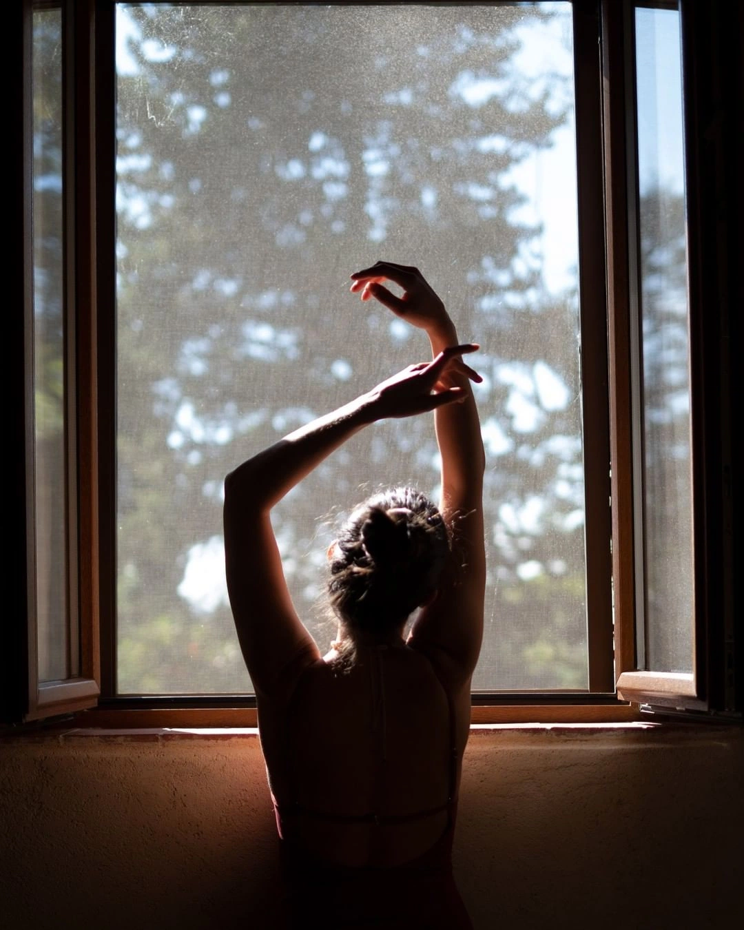 A woman seated before a tall sunlit window, arms raised in a slow, sensing gesture